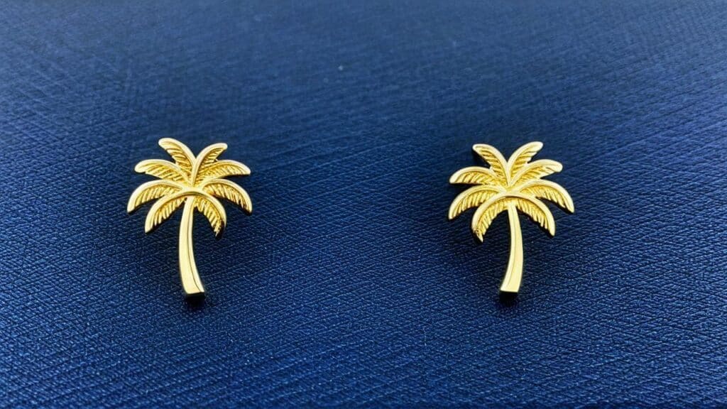 Pair of gold palm tree stud earrings with textured fronds on a navy blue fabric background.