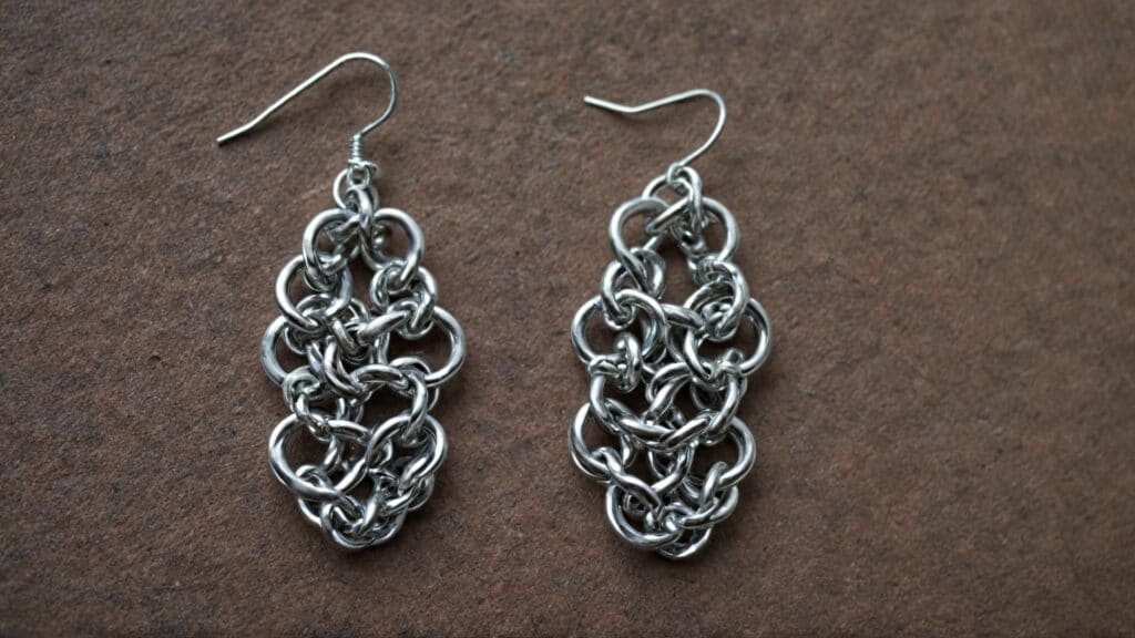 A set of silver-tone chainmaille earrings featuring a layered weave pattern, laid on a textured brown background.
