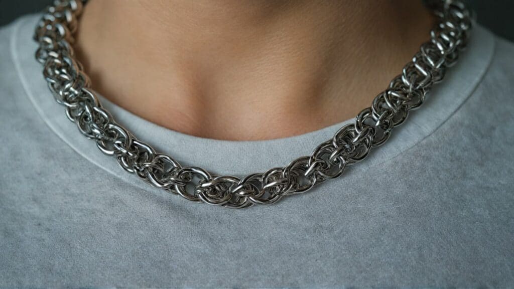 Close-up of a thick stainless steel chainmaille necklace worn over a light-colored shirt, showcasing intricate interwoven metal rings.