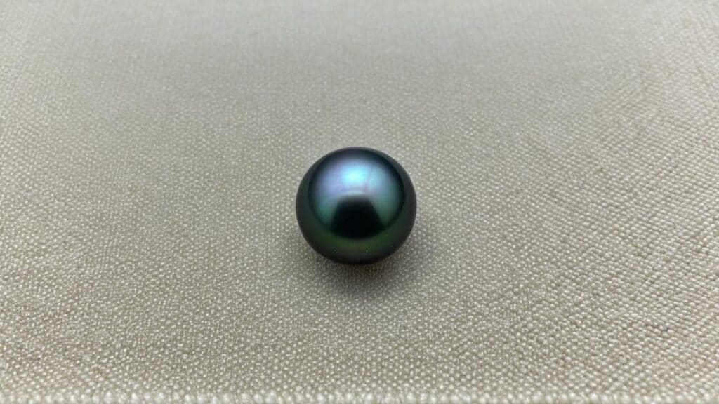 Close-up of a dark green Tahitian black pearl with subtle purple overtones placed on a textured fabric background.