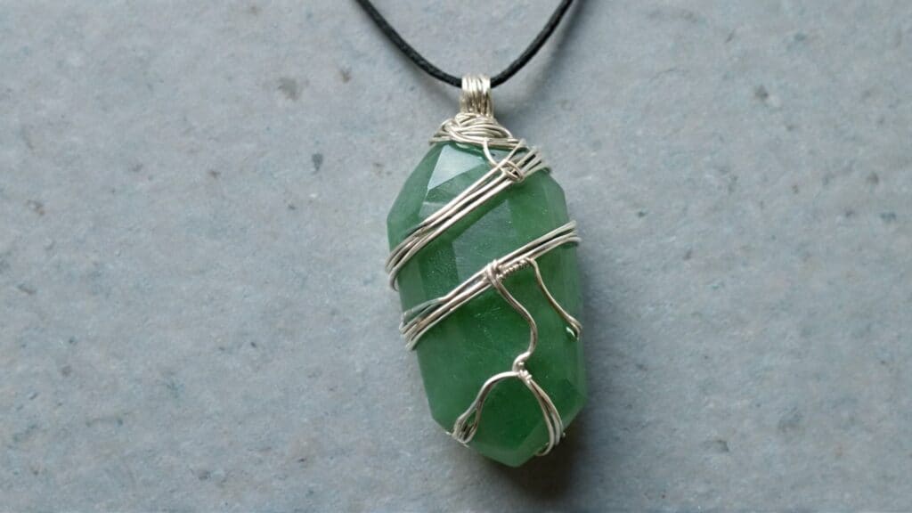 Close-up of a faceted green aventurine pendant wrapped in silver wire on a black cord, displayed on a gray background.