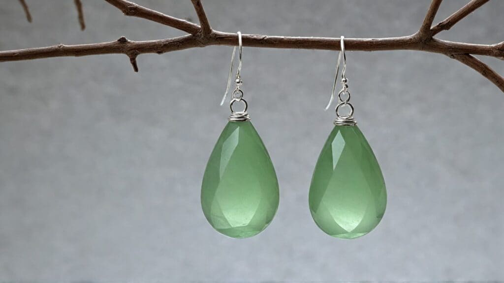 A pair of faceted green calcite teardrop earrings hanging from silver hooks on a twig, with a neutral gray background.