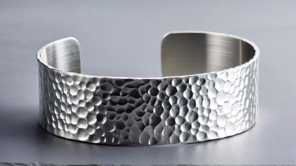 Hammered aluminum cuff bracelet with a textured finish, reflecting light on a sleek gray background.