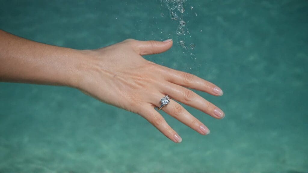 Woman's hand wearing diamond engagement ring submerged in clear water with bubbles, showing water exposure risks for jewelry