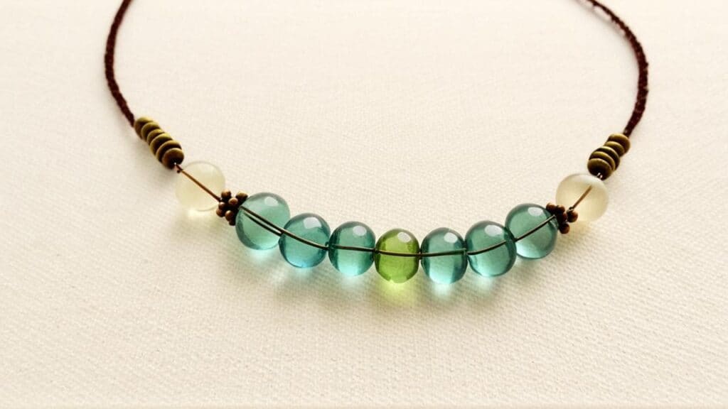 Handcrafted necklace featuring round glass beads in shades of teal, green, and white strung on a brown cord.