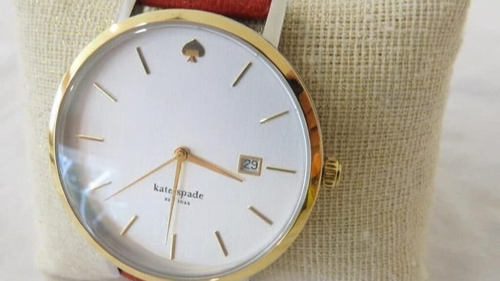 Kate Spade watch featuring gold-tone case, clean white dial with date window, signature lowercase branding, and red leather strap