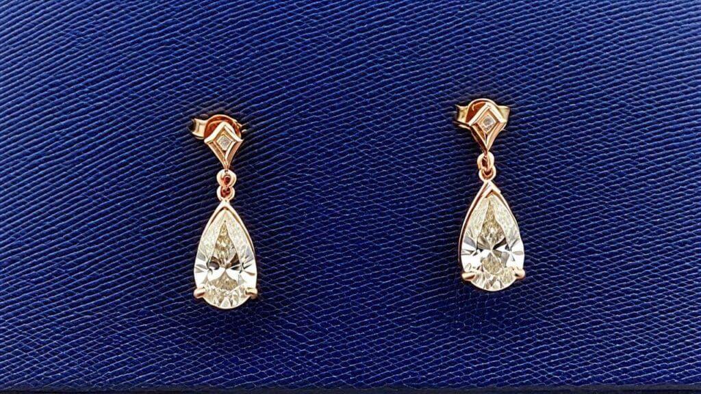 A pair of elegant pear-cut memorial diamond earrings set in gold, displayed on a textured blue background, symbolizing a timeless tribute to a loved one.