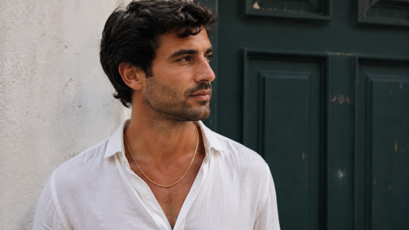 Man wearing a thin gold chain necklace with an open white linen shirt