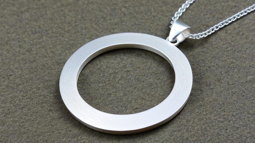 Close-up of a minimalist aluminum circle pendant necklace with a brushed finish on a silver chain, displayed on a textured surface.