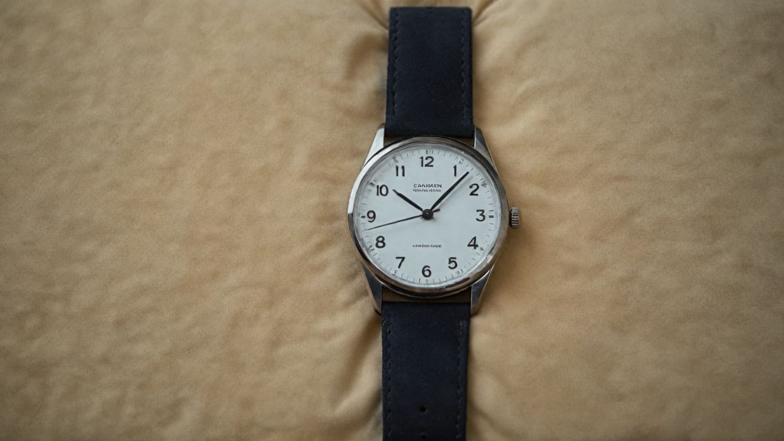 Steel watch with white numbered dial and navy strap on a beige cushion, no box