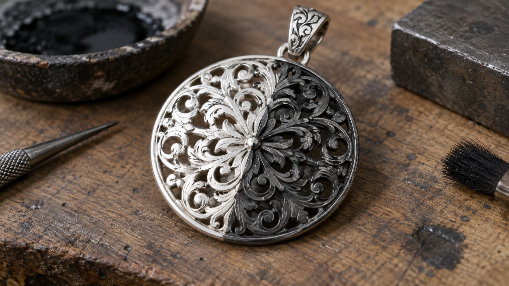 Close-up of an oxidized silver filigree pendant on a jeweler's workbench with liver of sulfur solution and tools