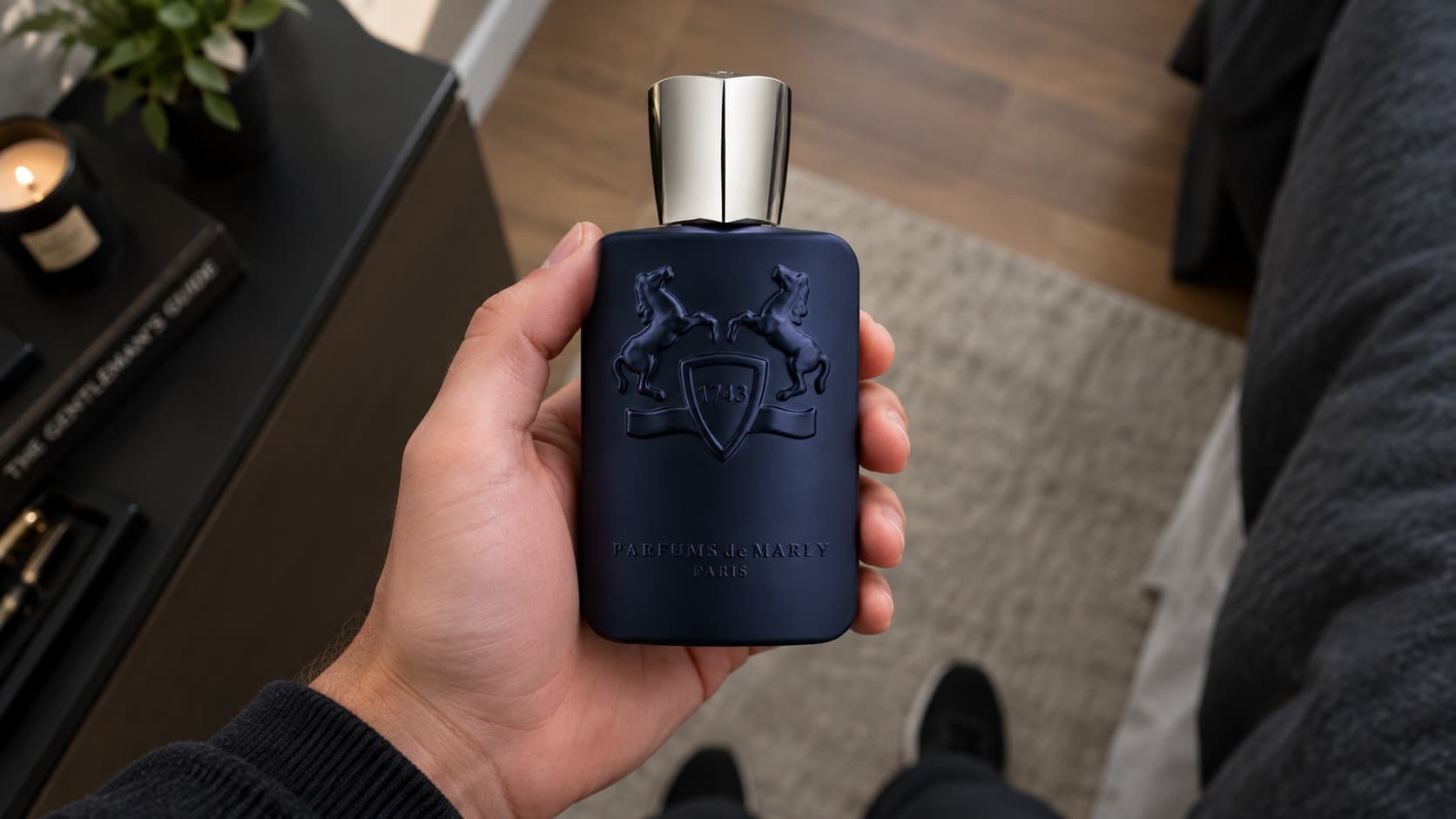 Point-of-view photo of a hand holding a Parfums de Marly Layton perfume bottle in a modern living room
