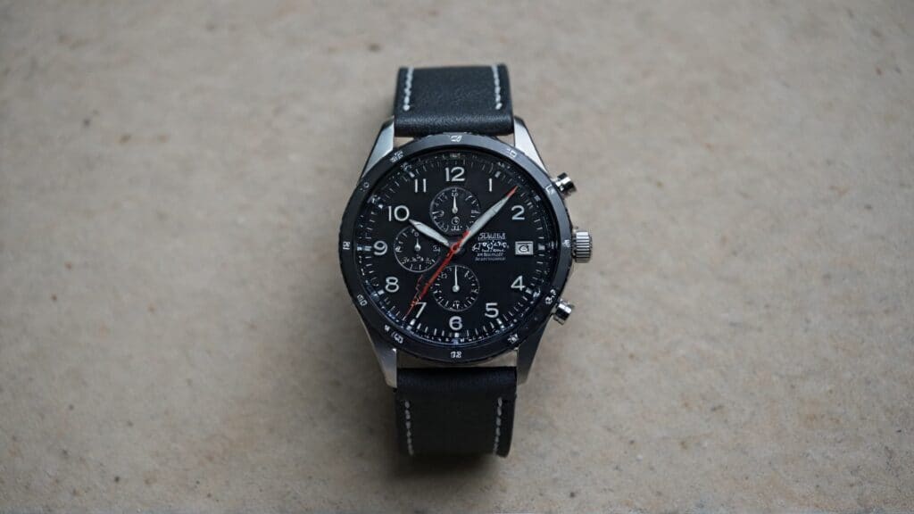 Racing chronograph watch with black dial, three subdials, tachymeter bezel, and black leather strap on concrete surface