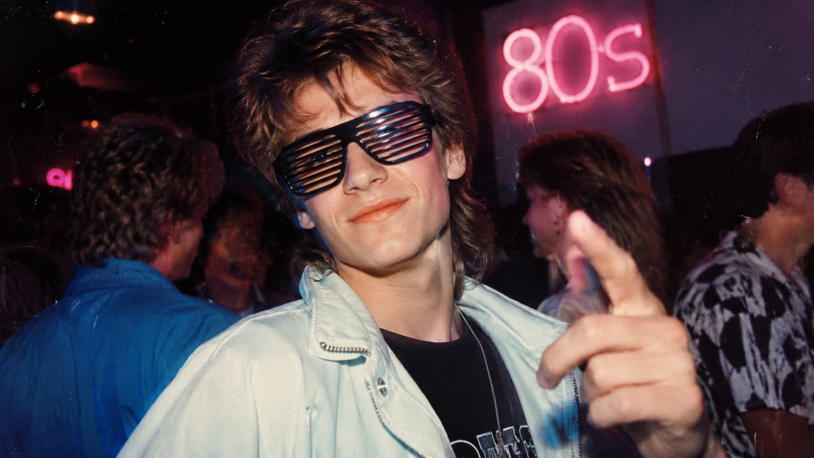 Person wearing black shutter shades sunglasses at an 80s-themed party with neon 80s sign