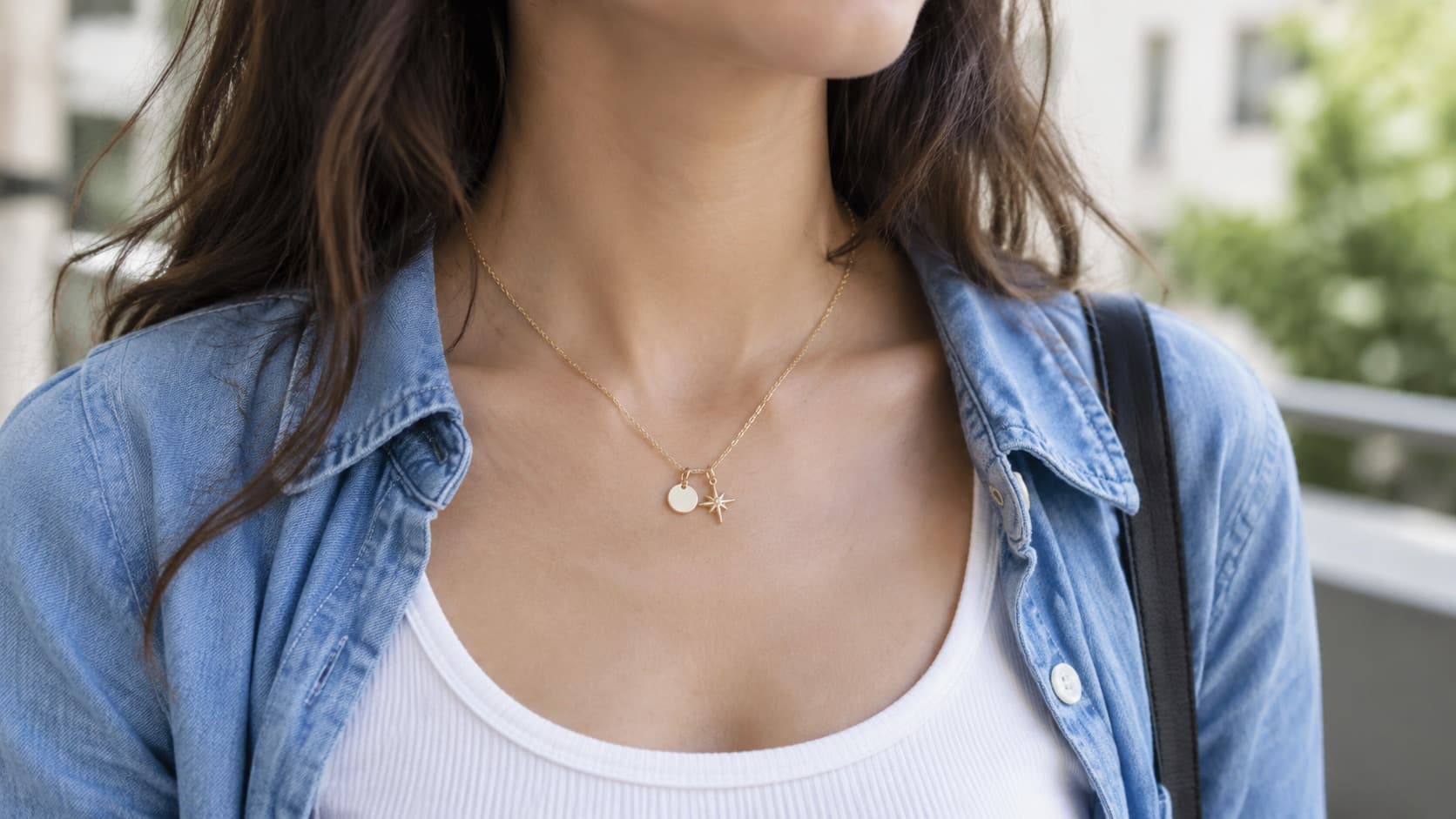 Close-up of a gold necklace with two staggered pendants, a disc charm and a star charm, worn over a denim shirt