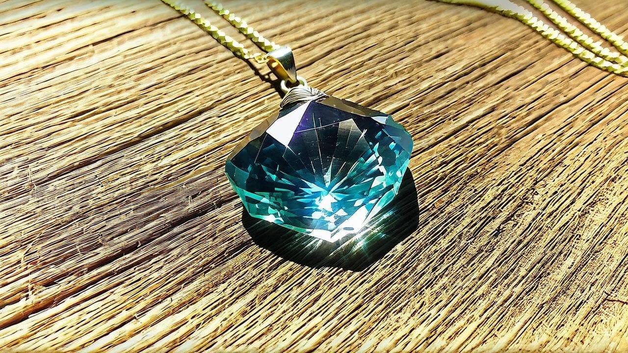 Faceted blue crystal pendant on a gold chain catching sunlight on woven wood