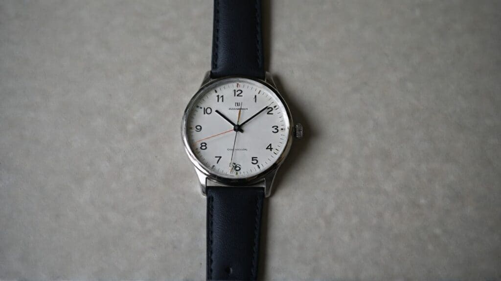 Clean Bauhaus watch featuring white dial, thin black hands, geometric hour markers, and minimal typography on navy leather strap