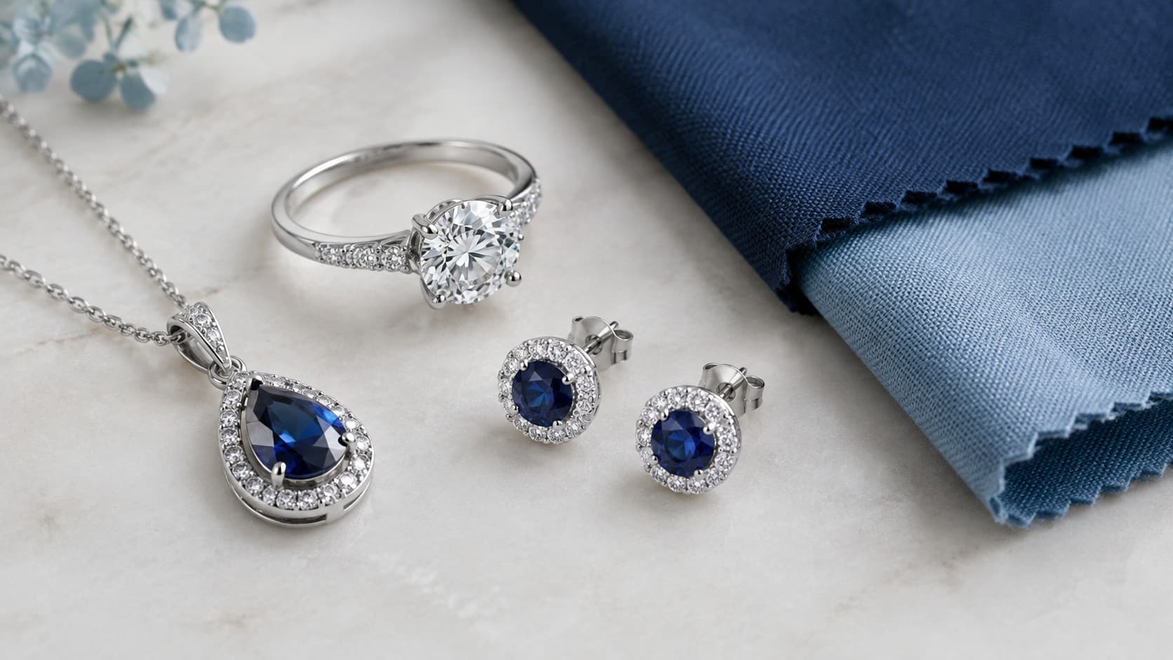 White gold necklace, ring, and stud earrings with sapphire stones displayed next to navy blue fabric swatches