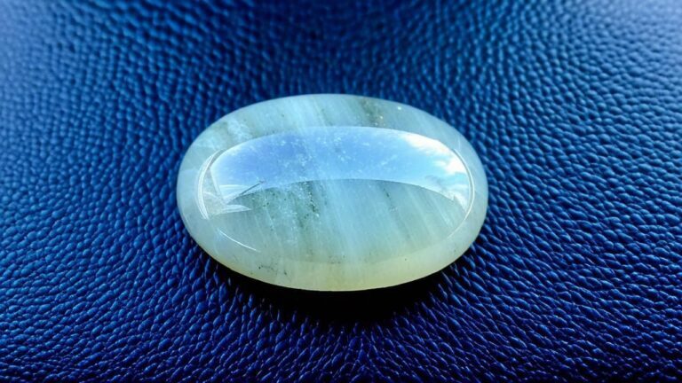 A cabochon-cut sillimanite gemstone with a silky luster and visible chatoyancy, resting on a blue background.